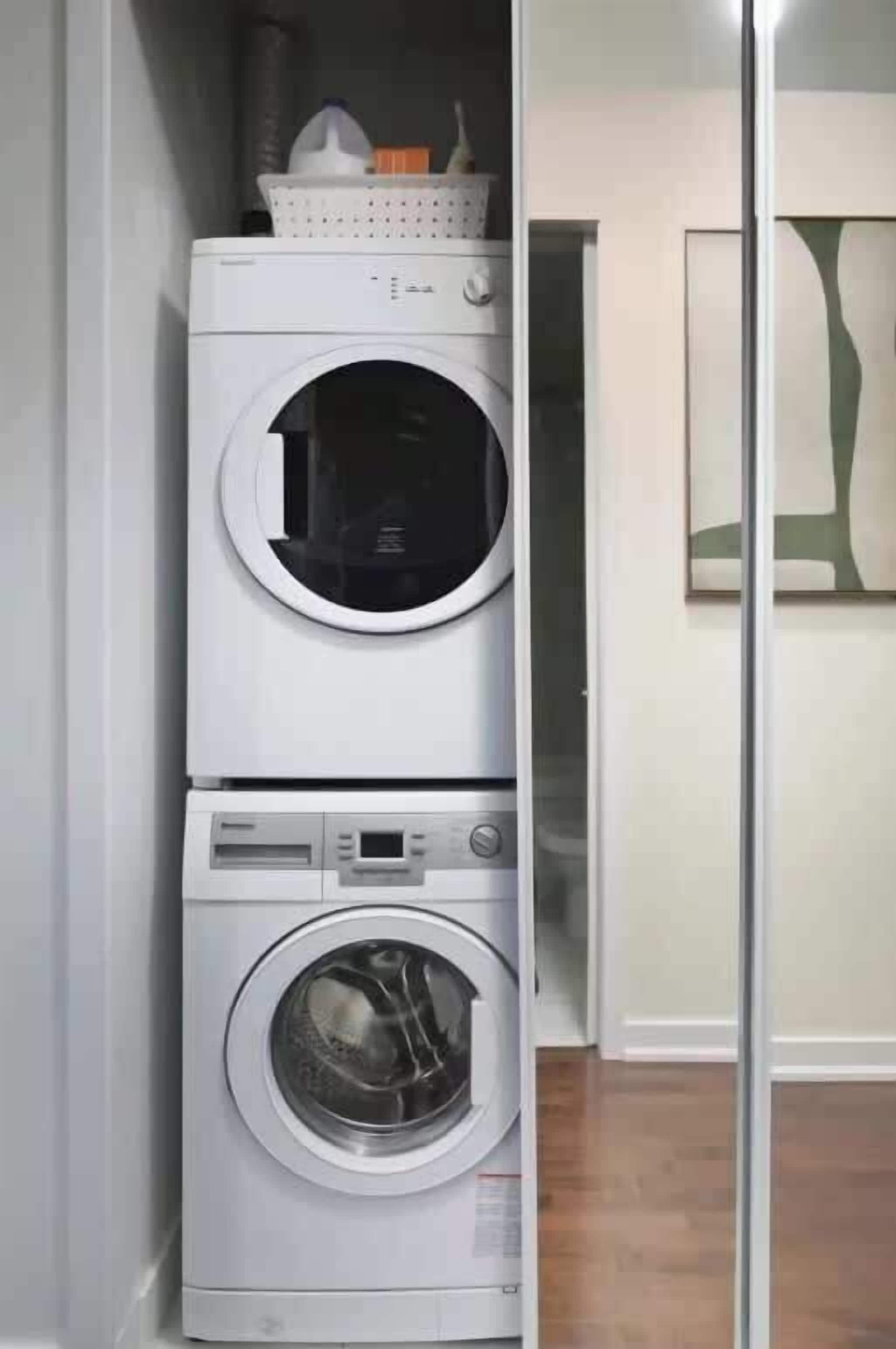 Une machine à laver et un sèche-linge superposés dans une buanderie, avec un plancher en bois et une œuvre d’art encadrée au mur.