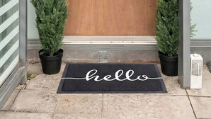 Een houten deur met een brievenbus, omlijst door twee potten met altijdgroene planten, en een 'welkom'-matje op de grond.