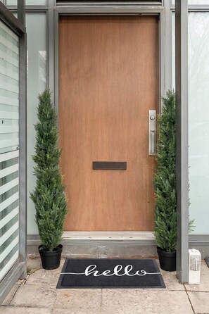 A wooden door with a mail slot, flanked by two potted evergreen plants, and a 'hello' mat on the ground. - Private Home, 3 BR, Rogers Centre & Free Parking (Toronto)