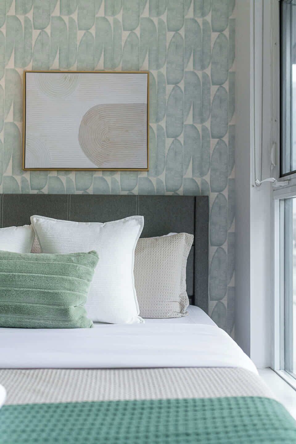 A neatly made bed with a grey headboard, white and green pillows, and a framed artwork above it.