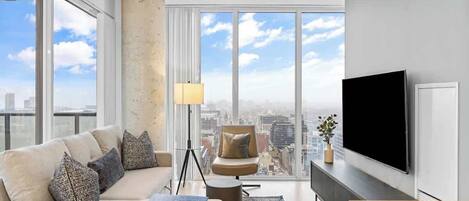 A modern living room with a large TV, a sofa set, a floor lamp, and a city view through large windows.