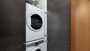A compact laundry room with a front-loading washer and dryer, a small shelf with cleaning supplies, and a wooden door. - Chic City Oasis with Cafes & Bars at your Doorstep! (Toronto)