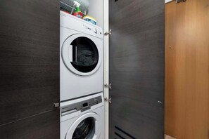 A compact laundry room with a front-loading washer and dryer, a small shelf with cleaning supplies, and a wooden door. - Chic City Oasis with Cafes & Bars at your Doorstep! (Toronto)
