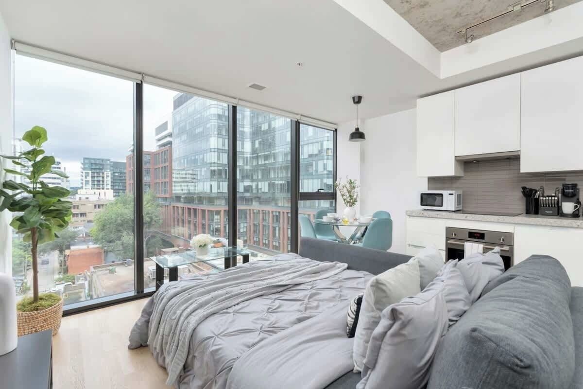 A modern bedroom with a large window, a bed, a dining area, and a kitchenette.