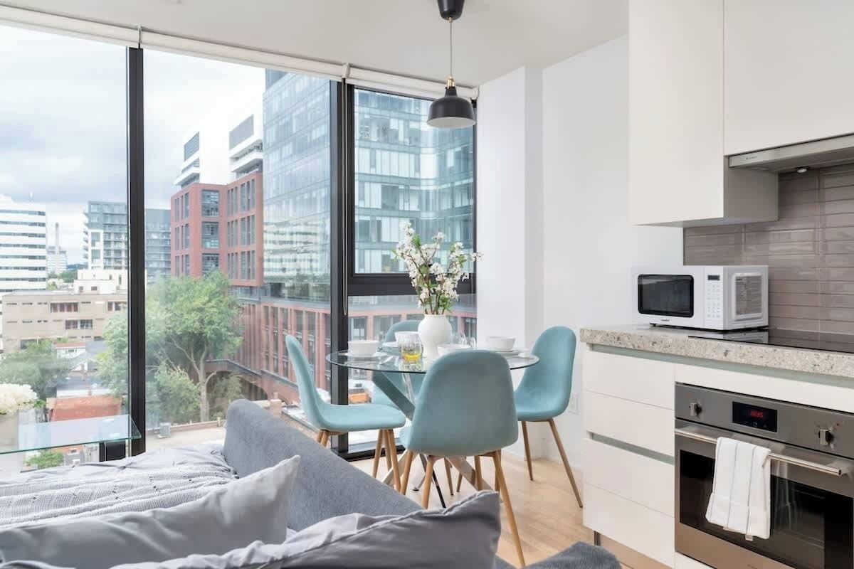A modern apartment with a large window, a dining area, a kitchenette, and a bed.