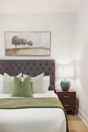 Ein Schlafzimmer mit einer gesteppten Kopfteil, einem Bett mit weißer Bettwäsche, grünen Kissen, einem Nachttisch mit Lampe und einem Bild über dem Bett.