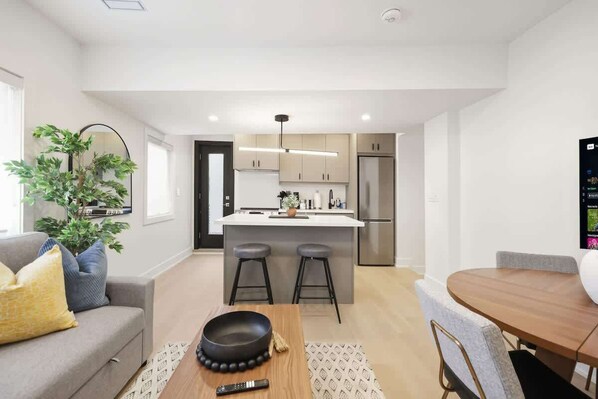 A modern kitchen with a central island, bar stools, a dining table with chairs, and a living area with a sofa and a plant. - Elegant 2BR Retreat, Sleek Design & Comfort | Parking Available (Toronto)