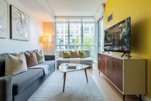 A modern living room with a sofa, a coffee table, a TV, and a window with a view of buildings. - 3BR/2BA City Views + Free Parking | Parking Available (Toronto)