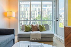 A modern living room with a grey sofa, a floor lamp, and a view of a building through large windows. - 3BR/2BA City Views + Free Parking | Parking Available (Toronto)