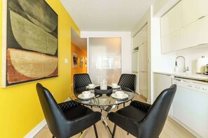 A dining area with a glass table and black chairs, a kitchen with white cabinets, and a yellow wall with a large abstract painting. - 3BR/2BA City Views + Free Parking | Parking Available (Toronto)
