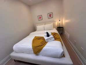 A neatly made bed with white linens and a mustard-colored throw blanket, a bedside lamp, and two framed artworks on the wall.
