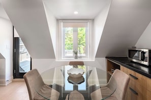 A modern kitchen with a glass dining table, two chairs, a microwave, and a window with a view of greenery.