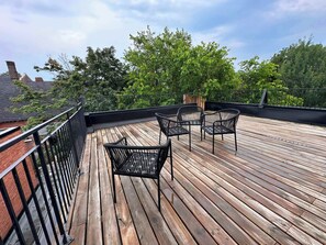 A wooden deck with metal railings, two wicker chairs, and a view of trees and buildings.