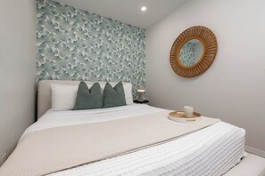 Un lit bien fait, recouvert d’une couverture blanche à texture intéressante, un mur décoratif orné d’un motif végétal, un miroir rond en fibres tressées et une table de chevet avec une lampe et un plateau.