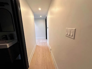A narrow hallway with a wooden floor, white walls, and a light switch on the right wall.
