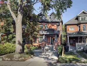 A residential building with a brick facade, a prominent entrance with steps, and a large tree in the foreground.