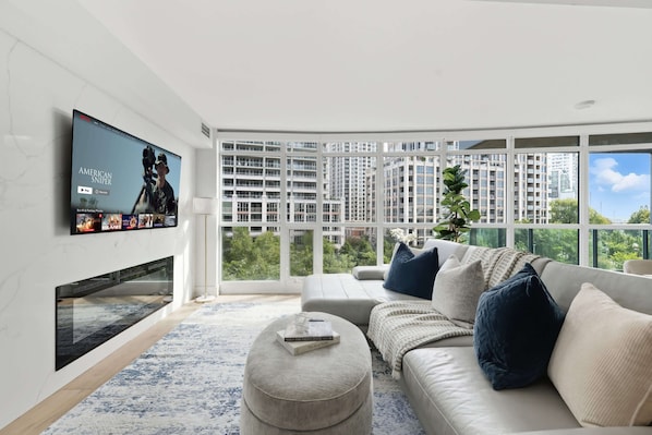 A modern living room with a flat-screen TV, a sofa, and a large window offering a city view. - Upscale 2BR Condo Downtown TO Gem w Parking | Parking Available (Toronto)