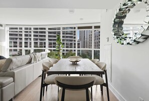 A modern living room with a dining table, sofa, and large windows offering a view of high-rise buildings. - Upscale 2BR Condo Downtown TO Gem w Parking | Parking Available (Toronto)