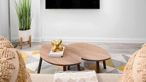 A modern living room with a flat-screen TV mounted on the wall, two wooden coffee tables, a potted plant, and a patterned rug.