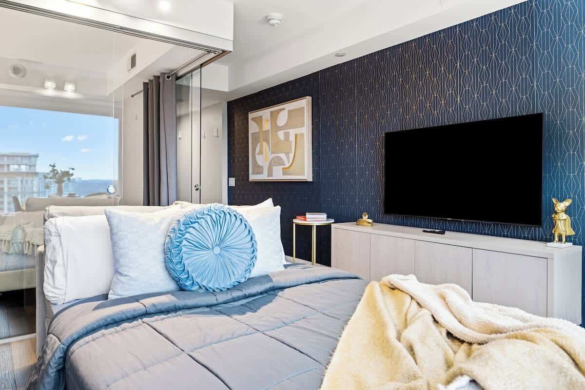 A modern bedroom with a large bed, a flat-screen TV, and a city view.