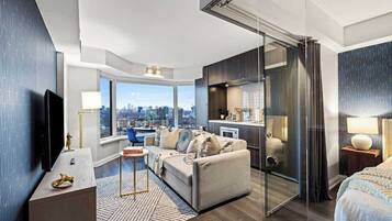 A modern hotel room with a city view, a sofa, a dining table, and a kitchen area.