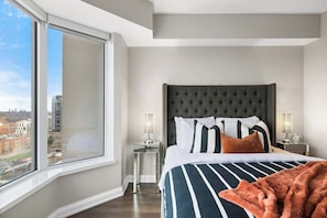 A hotel room with a large window, a tufted headboard, a striped bedspread, and two bedside lamps.