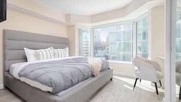 A modern bedroom with a large bed, a seating area, and a view of the city through large windows.
