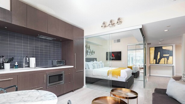 A modern hotel room with a kitchen area, a bed with a glass partition, and a dining area with a round table. - Designer 4 Seasons Luxury Bldg of Yorkville! (Toronto)
