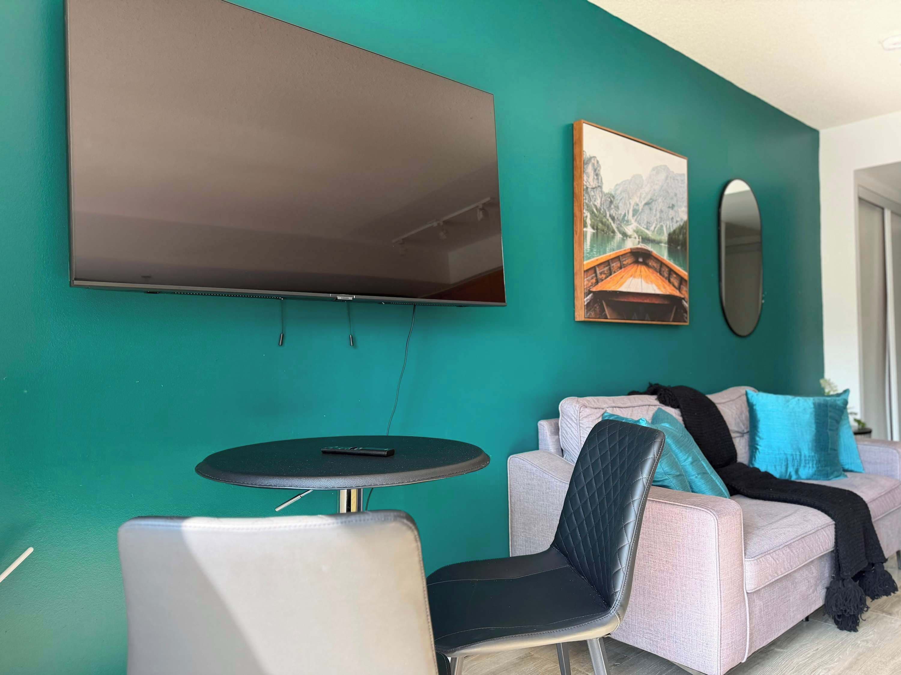 Un mur de couleur turquoise, sur lequel est fixé un téléviseur à écran plat au-dessus d’une table ronde et de deux chaises. Un canapé avec des coussins est placé contre le mur, et un tableau encadré est accroché au-dessus.