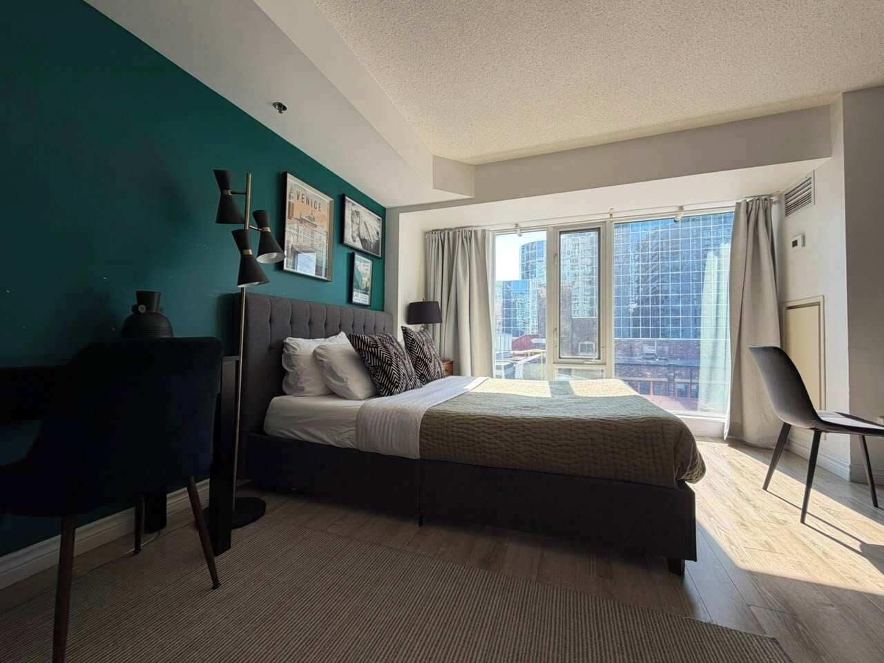 A modern bedroom with a large bed, a desk, and a view of the city through a large window.