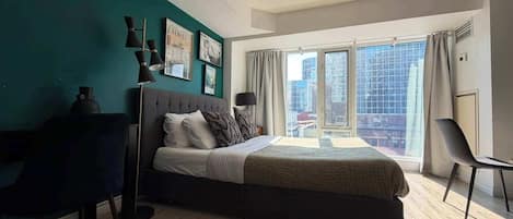 Un dormitorio moderno con una cama grande, un escritorio y vistas de la ciudad a través de un ventanal amplio.