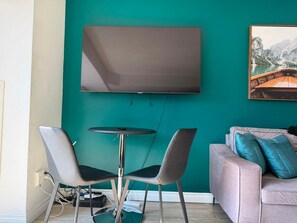 A modern living room with a teal wall, a flat-screen TV mounted above a round table, two chairs, and a sofa with cushions. - Modern Studio by Massey Hall | Heart of Toronto | Parking Available (Toronto)