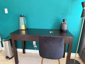 A teal-colored wall, a dark wooden table, a black chair with wooden legs, a trash bin, a floor lamp, and a vase. - Modern Studio by Massey Hall | Heart of Toronto | Parking Available (Toronto)