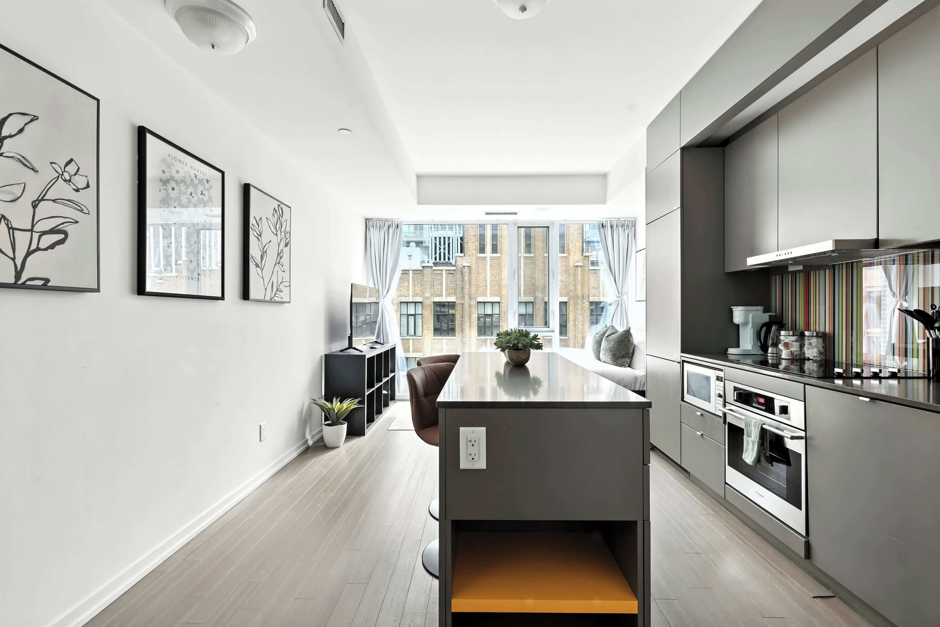 A modern kitchen with a central island, a dining area with a sofa and table, and large windows offering a view of the cityscape.