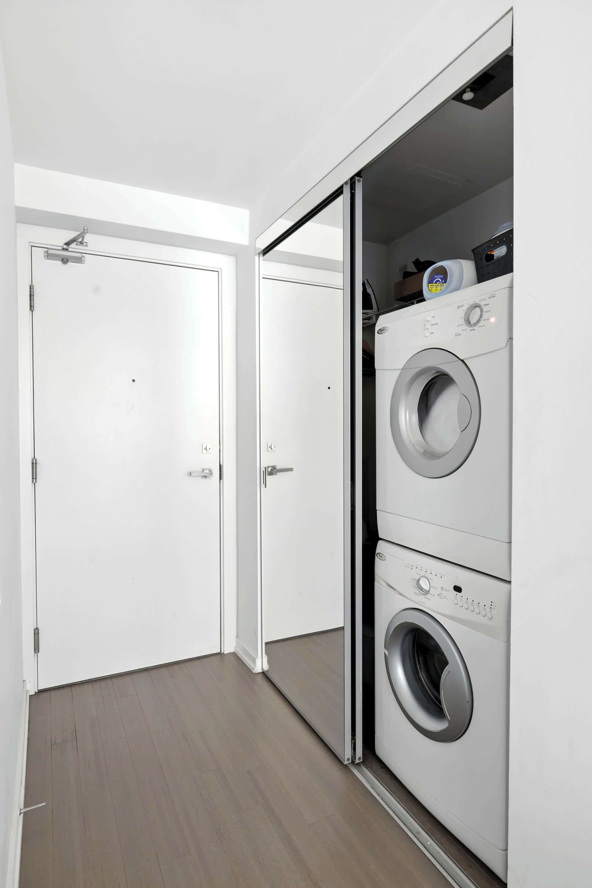 A laundry room with a front-loading washer and dryer, a white door, and a wooden floor.