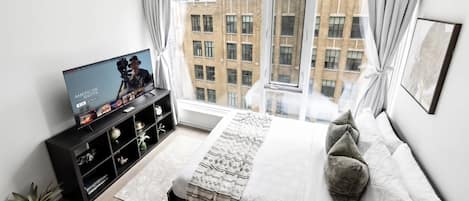 Quarto moderno com uma janela grande, televisão, cama com almofadas e uma pequena planta.