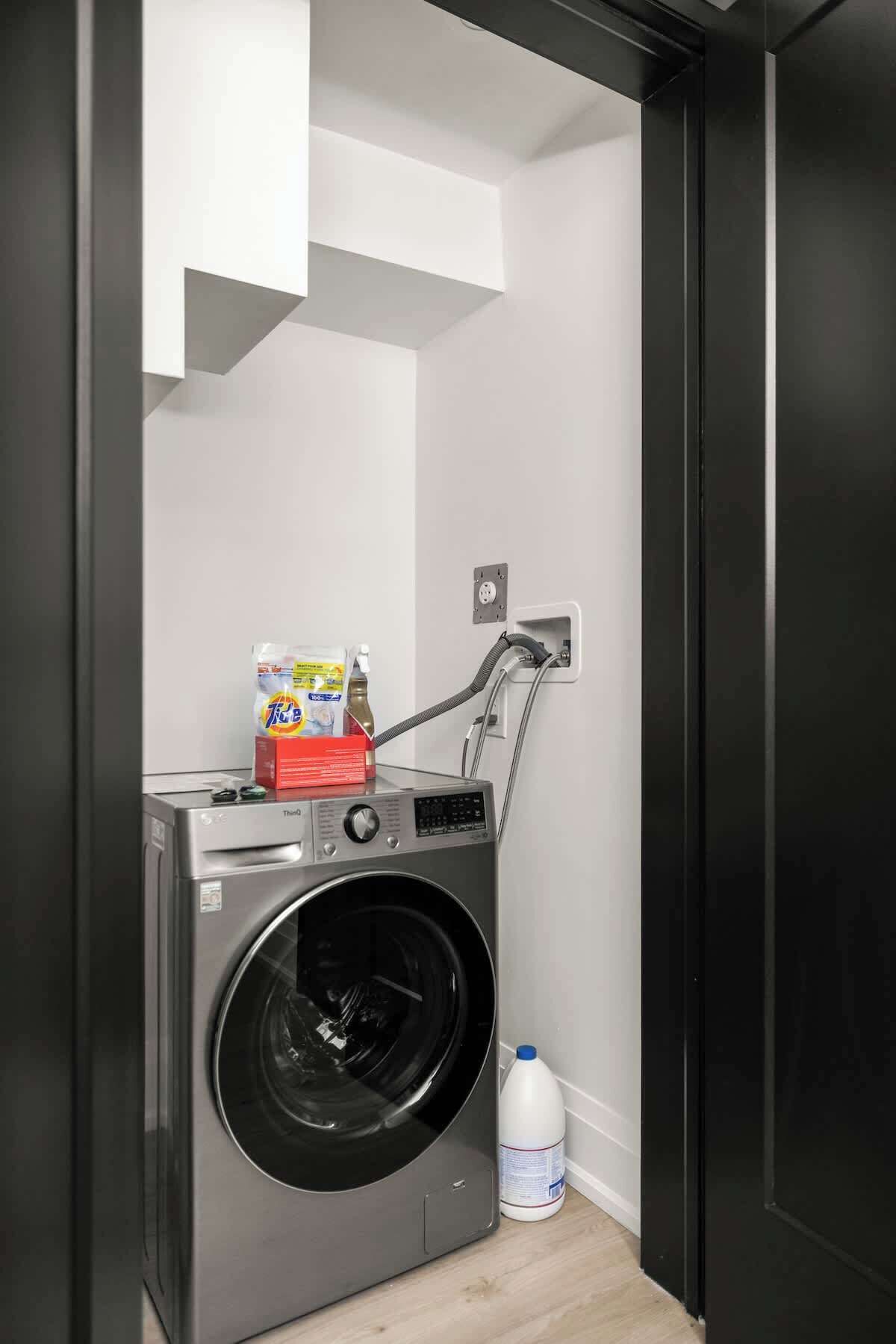 A compact laundry room with a front-loading washing machine, a detergent bottle, and a water jug.