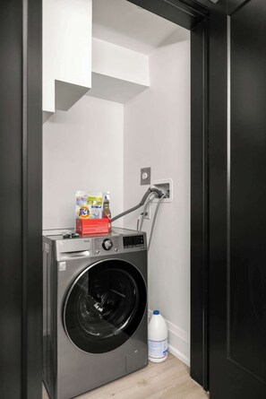 A compact laundry room with a front-loading washing machine, a detergent bottle, and a water jug.