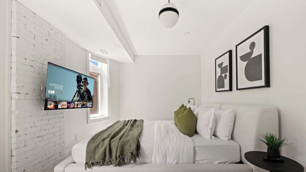 Un dormitorio moderno con pared de ladrillo blanco, un televisor de pantalla plana sobre la cama, un sofá blanco y una mesita auxiliar con una planta.