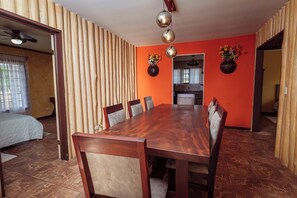 A dining area with a wooden table and chairs, a kitchen in the background, and a window with curtains. - The Teak House (San Rafael)