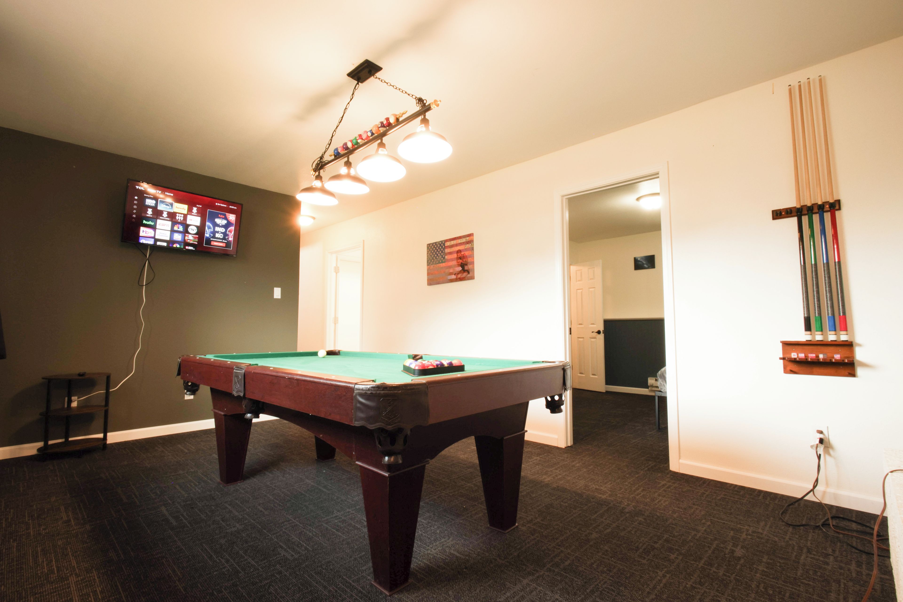 Une table de billard se trouve dans une pièce où un téléviseur est fixé au mur, un drapeau est accroché au mur et une porte donne accès à une autre pièce.