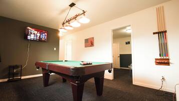 Une table de billard se trouve dans une pièce où un téléviseur est fixé au mur, un drapeau est accroché au mur et une porte donne accès à une autre pièce.