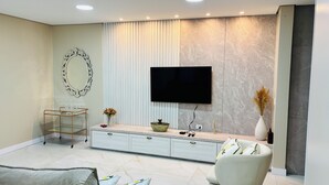Uma sala de estar moderna com uma televisão de tela plana na parede, uma mesa de centro de vidro, um sofá branco e um espelho decorativo.