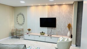 Ein modernes Wohnzimmer mit einem wandmontierten Flachbildfernseher, einem Glastisch, einem weißen Sofa und einem dekorativen Spiegel.