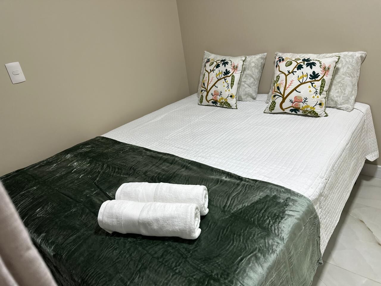 Een net opgemaakt bed met een groene spreideken en twee decoratieve kussens, een witte handdoek en een witte muur met een lichtschakelaar.