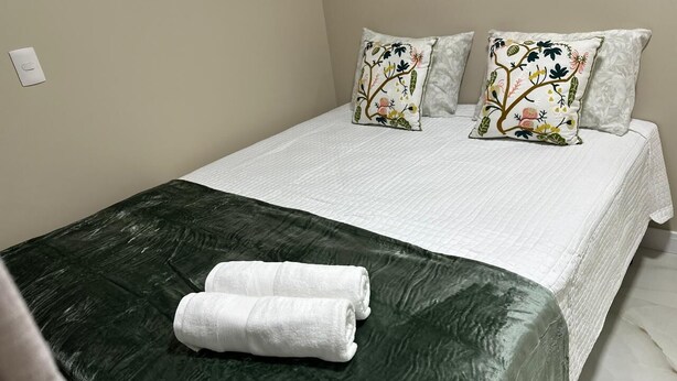 Uma cama bem arrumada com um edredom verde e duas almofadas decorativas, uma toalha branca e uma parede clara com um interruptor de luz.