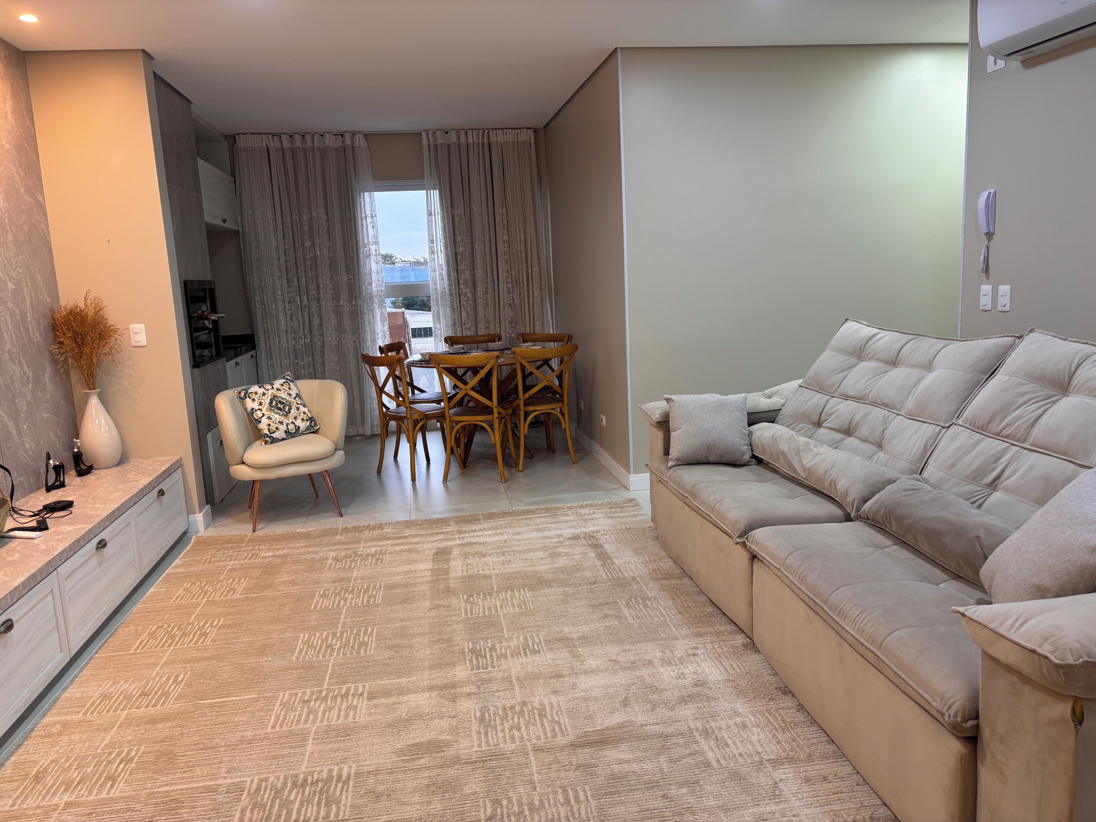 Uma sala de estar com um sofá, uma mesa de jantar com cadeiras, uma mesinha com um vaso e um telefone fixado na parede.