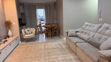 Uma sala de estar com um sofá, uma mesa de jantar com cadeiras, uma mesinha com um vaso e um telefone fixado na parede.