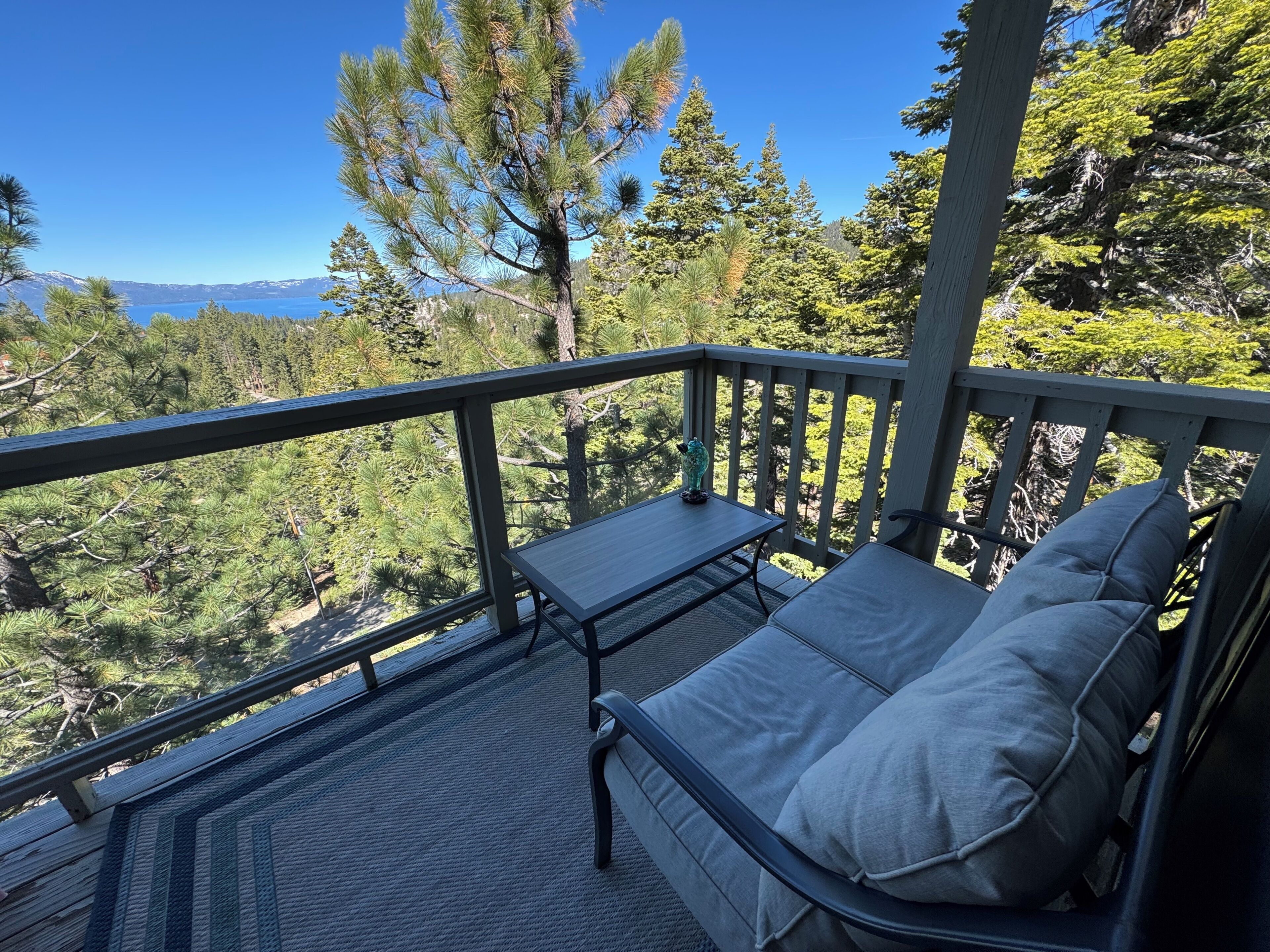 A balcony with a table and seating, overlooking a forested area and a lake.