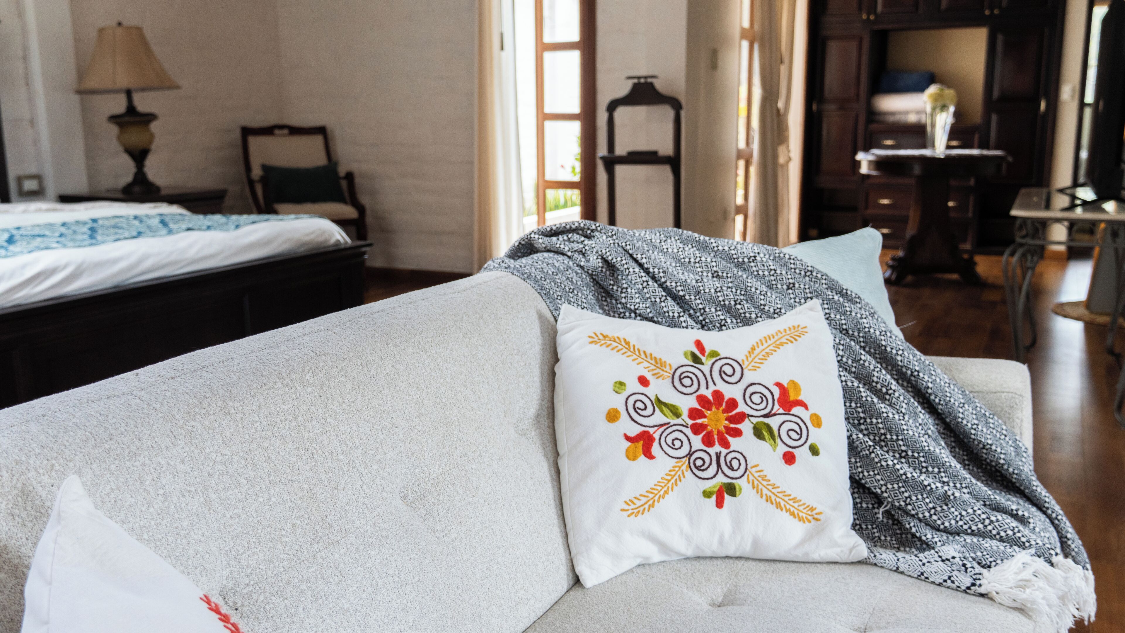 A neatly made bed with a patterned pillow, a bedside table with a lamp, and a view of a dining area.